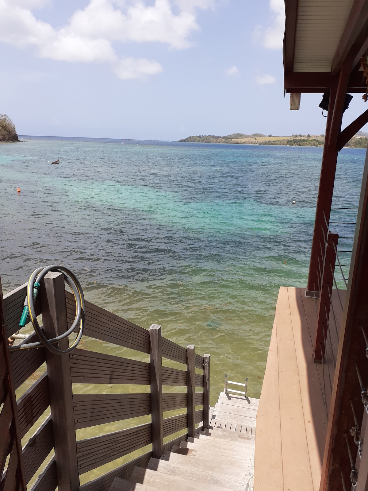 A staircase leads down to the water's edge, with a view of the clear blue sea. Gentle waves lap against the shore, and distant green hills are visible in the background under a partly cloudy sky.