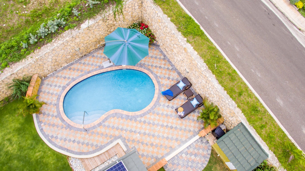 An aerial view of a private swimming pool is showcased, surrounded by patterned tiles and lush greenery. Two lounge chairs, one in blue and one in brown, are positioned next to the pool. A large green umbrella provides shade, while a neatly manicured lawn complements the setting.