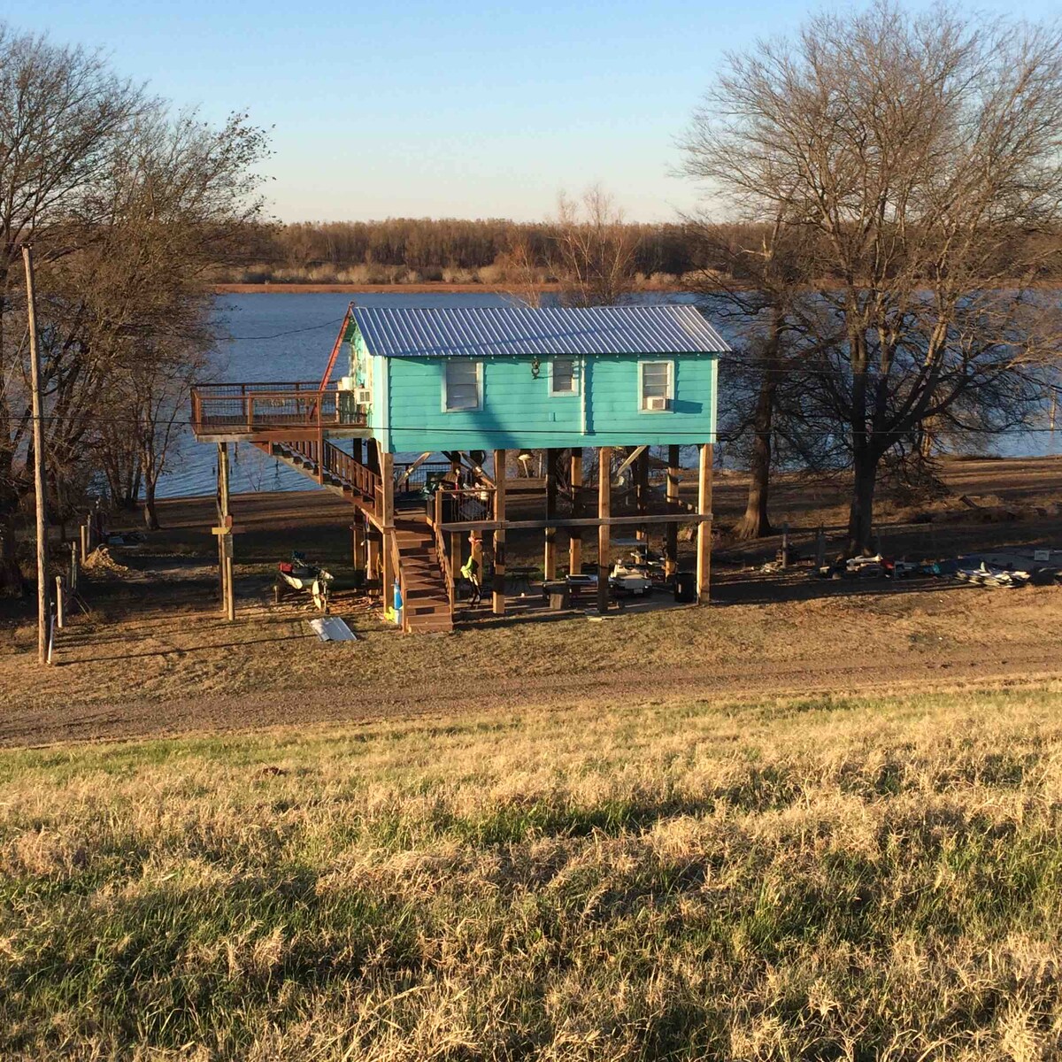A cabin with a bright blue exterior is elevated on stilts beside the water, surrounded by trees. Steps lead up to the entrance, and recreational equipment is visible on the ground below. The serene landscape includes a calm lake in the background.