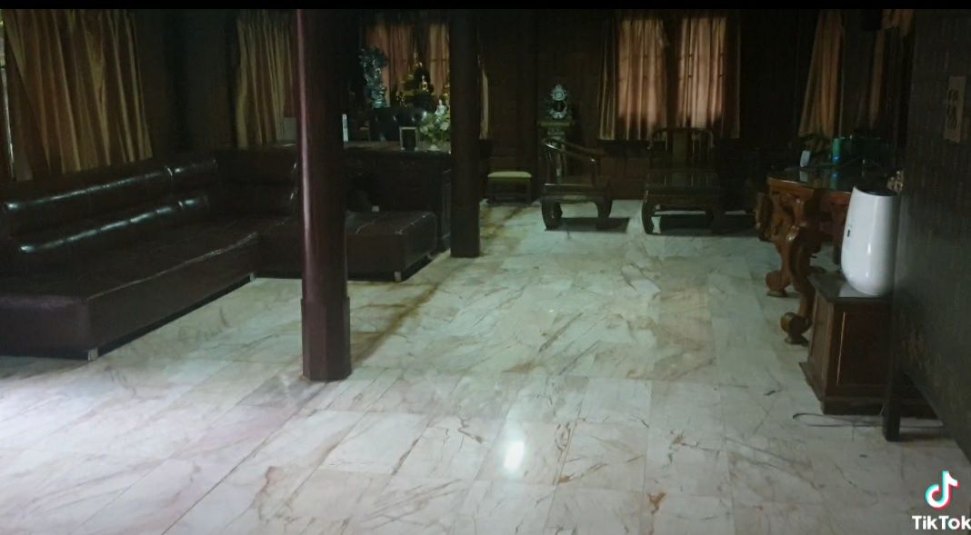 The living area features a spacious layout with polished marble flooring. A dark leather sectional sofa is positioned against one wall, while additional wooden furniture occupies the space. Natural light filters through large curtains, enhancing the traditional ambiance.