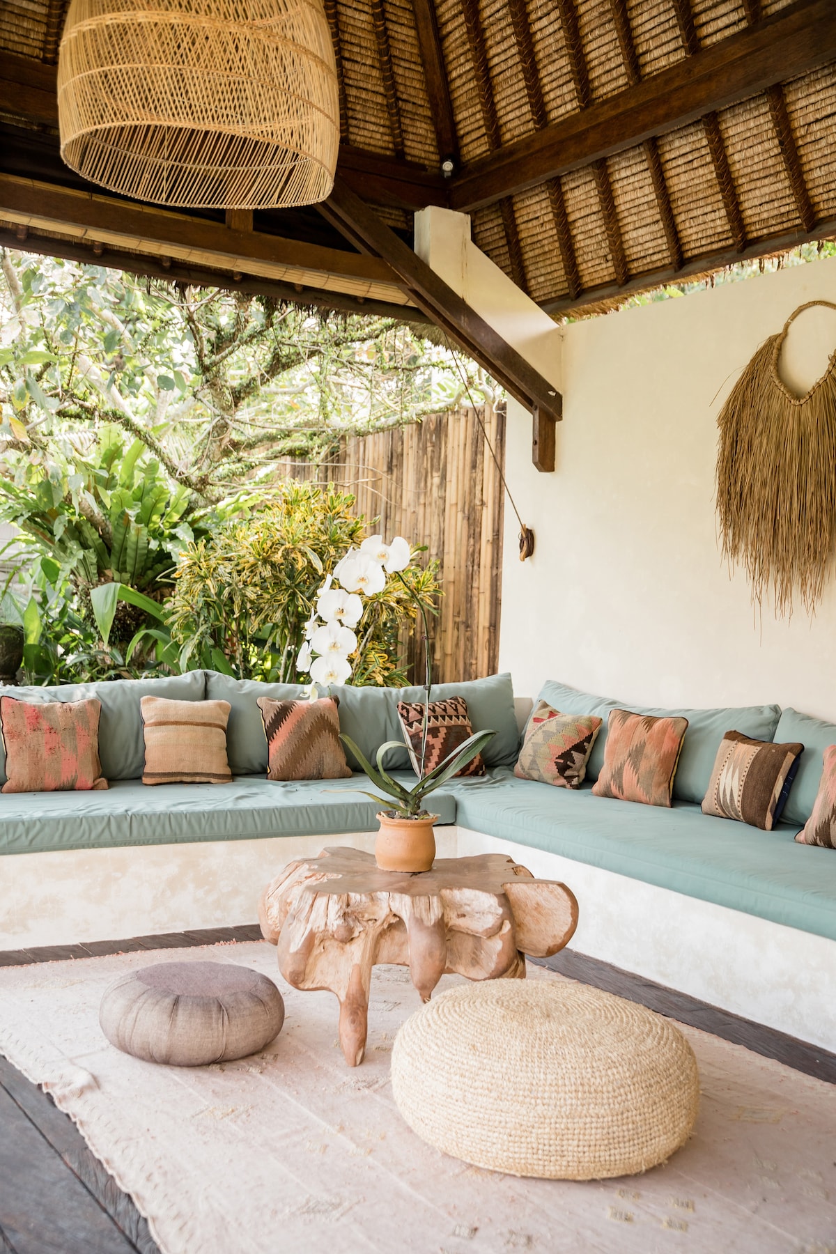 A spacious outdoor lounge area features a curved seating arrangement with decorative cushions in earthy tones. A rustic wooden table sits at the center, adorned with a small potted plant. Natural textures are highlighted by the woven light fixture and surrounding tropical greenery.