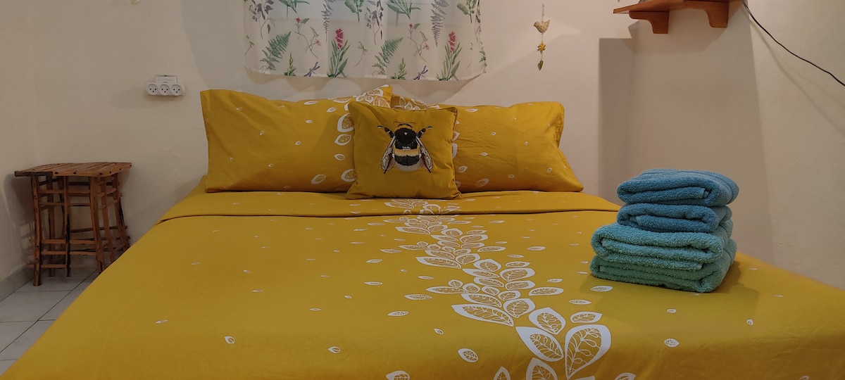 A cozy sleeping area is presented with a vibrant yellow bedspread, adorned with a floral pattern. A decorative bee pillow adds a personal touch. Neatly folded towels in turquoise are arranged beside the bed. Soft light enters through a window with a floral curtain.