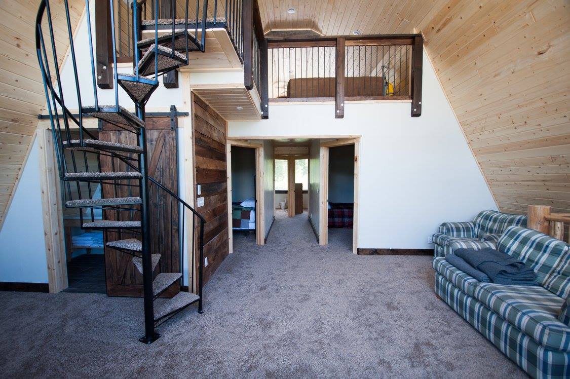 An open loft area features a spiral staircase leading to additional private bedrooms. Soft carpeting covers the floor, while natural wood walls create a warm atmosphere. A plaid sofa is positioned against one wall, providing seating and a cozy spot to relax.