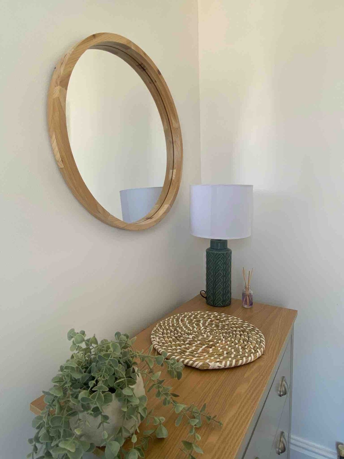 A round wooden mirror is mounted on a light-colored wall, reflecting the soft light in the room. Below it, a wooden side table is adorned with a textured placemat and a green ceramic lamp, accompanied by a decorative plant in a neutral pot.