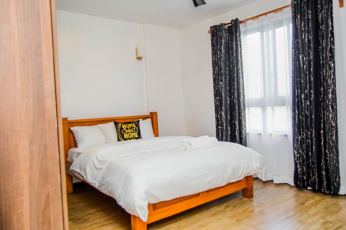 A spacious bedroom is featured with a wooden bed frame holding a white comforter and pillows. A decorative cushion reading 'Home Sweet Home' is positioned on the bed. Natural light filters through a large window, adorned with dark, patterned curtains.