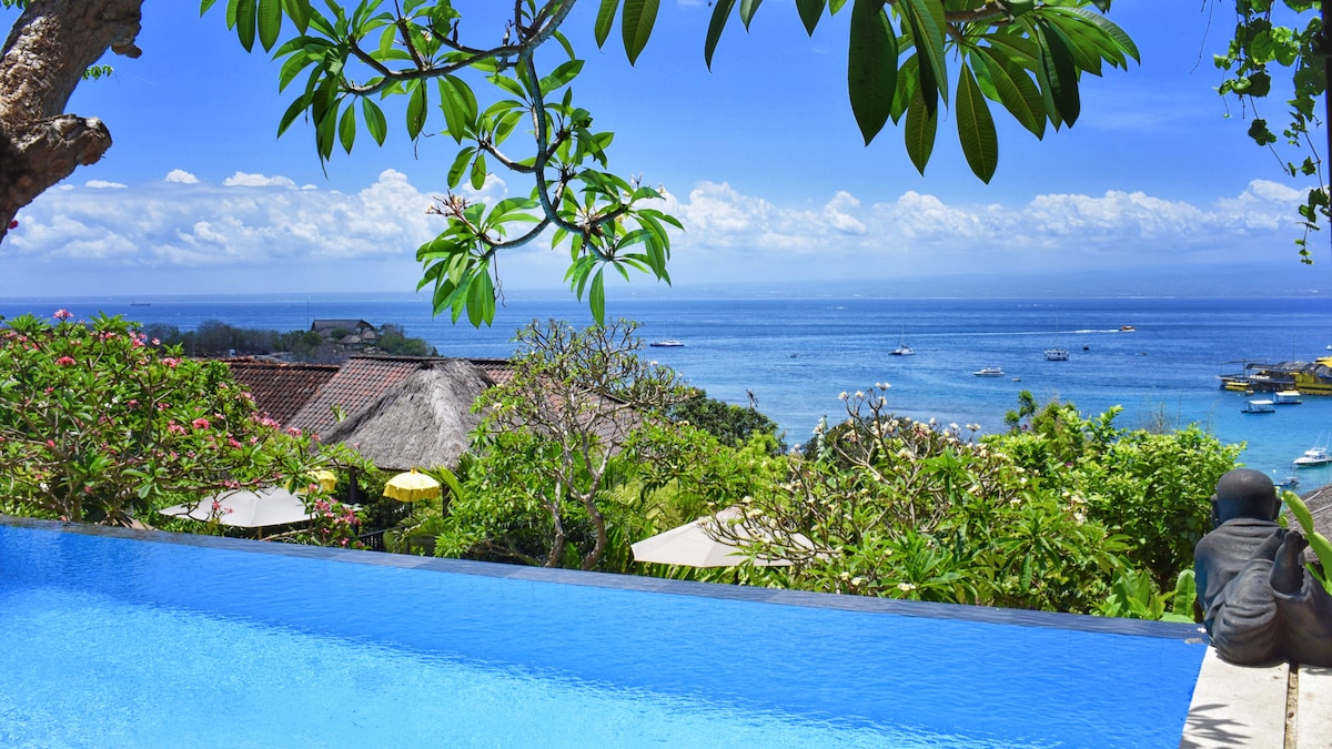 An infinity pool is seen overlooking the vibrant blue sea, complemented by lush greenery and tropical trees. Boats are anchored in the clear water, with distant landforms visible under a bright sky dotted with fluffy clouds.