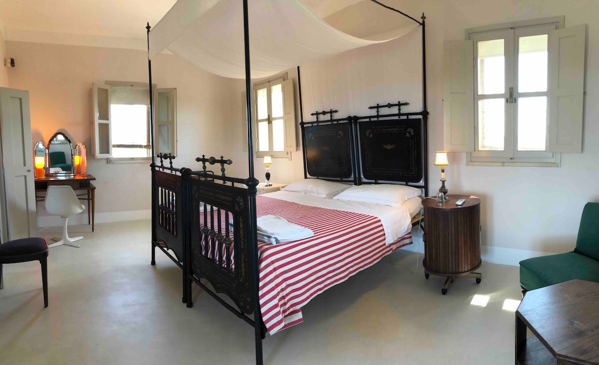 A spacious bedroom is showcased, featuring a spacious bed with a striped red and white coverlet. Large windows are adorned with shutters that allow natural light to enter. A desk with a mirror and a comfortable chair are positioned in the corner, along with a cozy seating area.