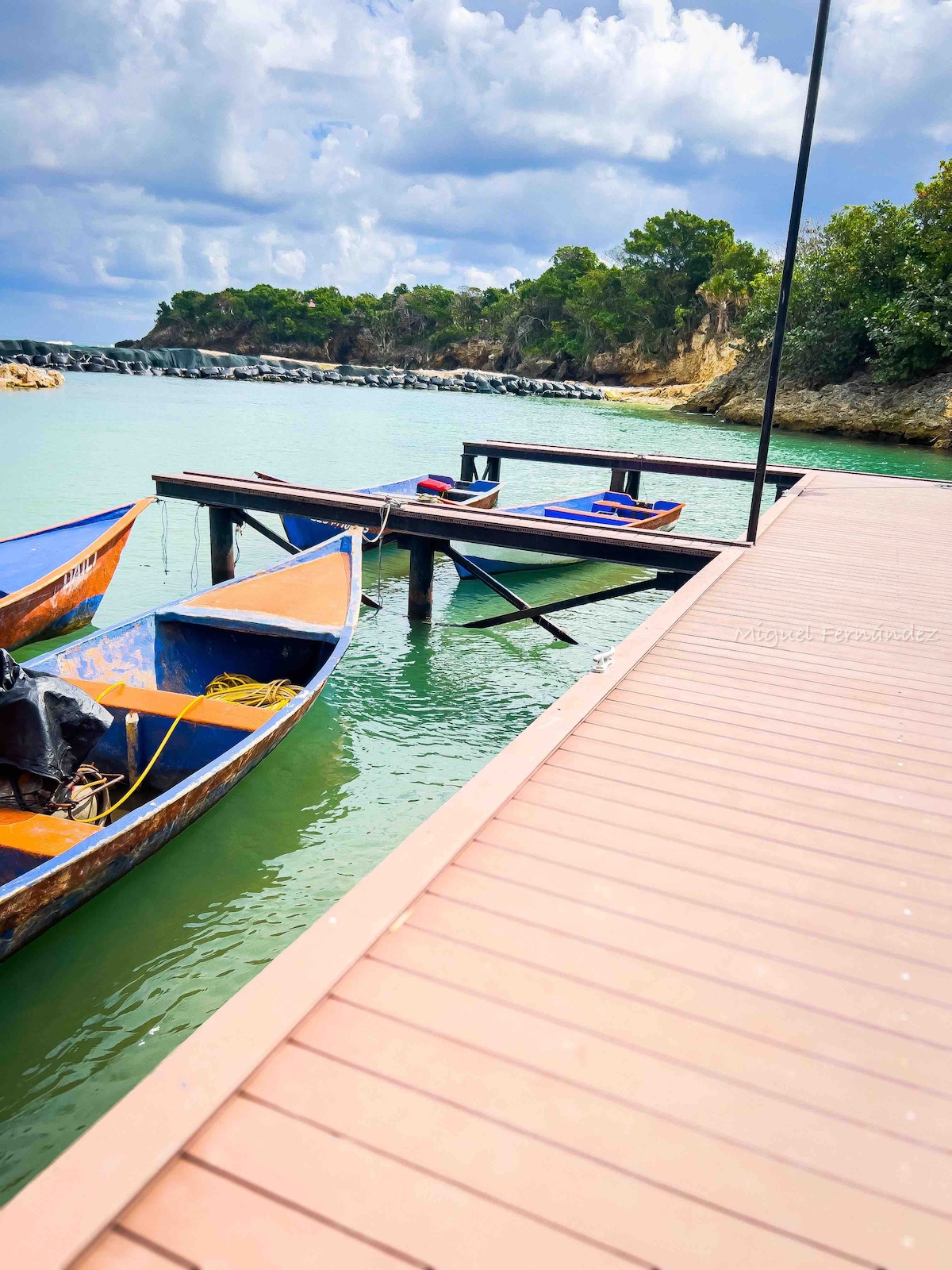 A wooden dock extends over calm waters, featuring several small fishing boats in vibrant colors. Lush greenery rises along the shore, while a rocky breakwater is visible in the distance under a partly cloudy sky.