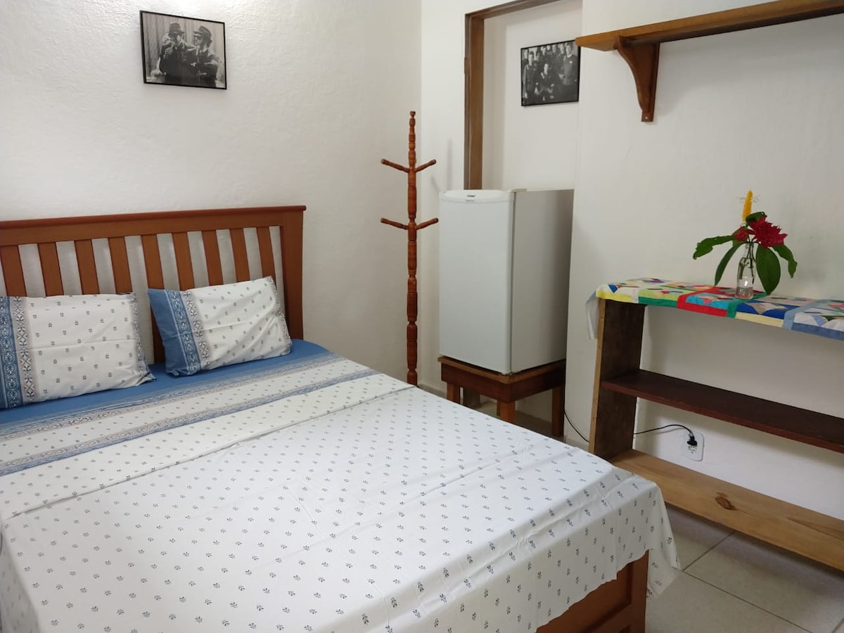 A cozy sleeping area features a spacious bed adorned with a patterned white and blue bedspread. A white refrigerator is positioned against the wall, while a small shelf displays a floral arrangement. A wooden coat rack is also visible, adding functionality to the space.