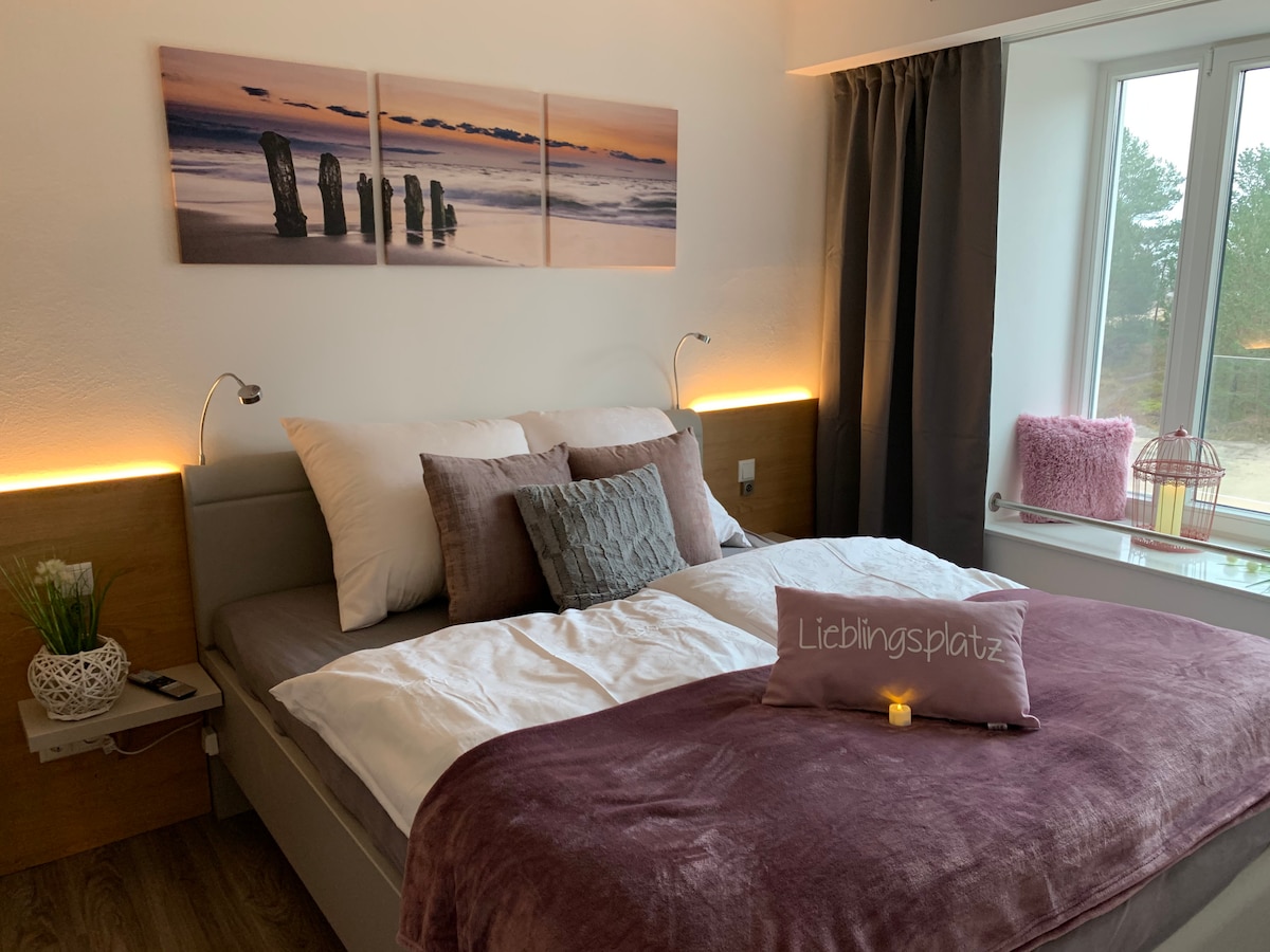 A spacious bed is adorned with neutral and pastel-colored pillows, complemented by a soft purple throw blanket. Above the bed, a triptych photograph depicts a tranquil coastal scene. Natural light enters through a large window, framed by gray curtains and partially revealing a glimpse of the outdoors.