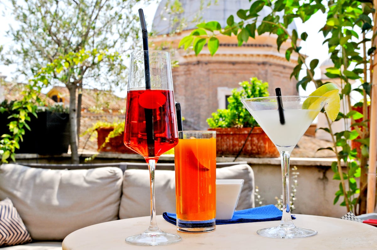 Three colorful cocktails are presented on a light-colored table. A vibrant red drink with a cherry, an orange drink, and a clear cocktail garnished with a lemon slice. Lush greenery and a distant historic dome provide a serene backdrop.