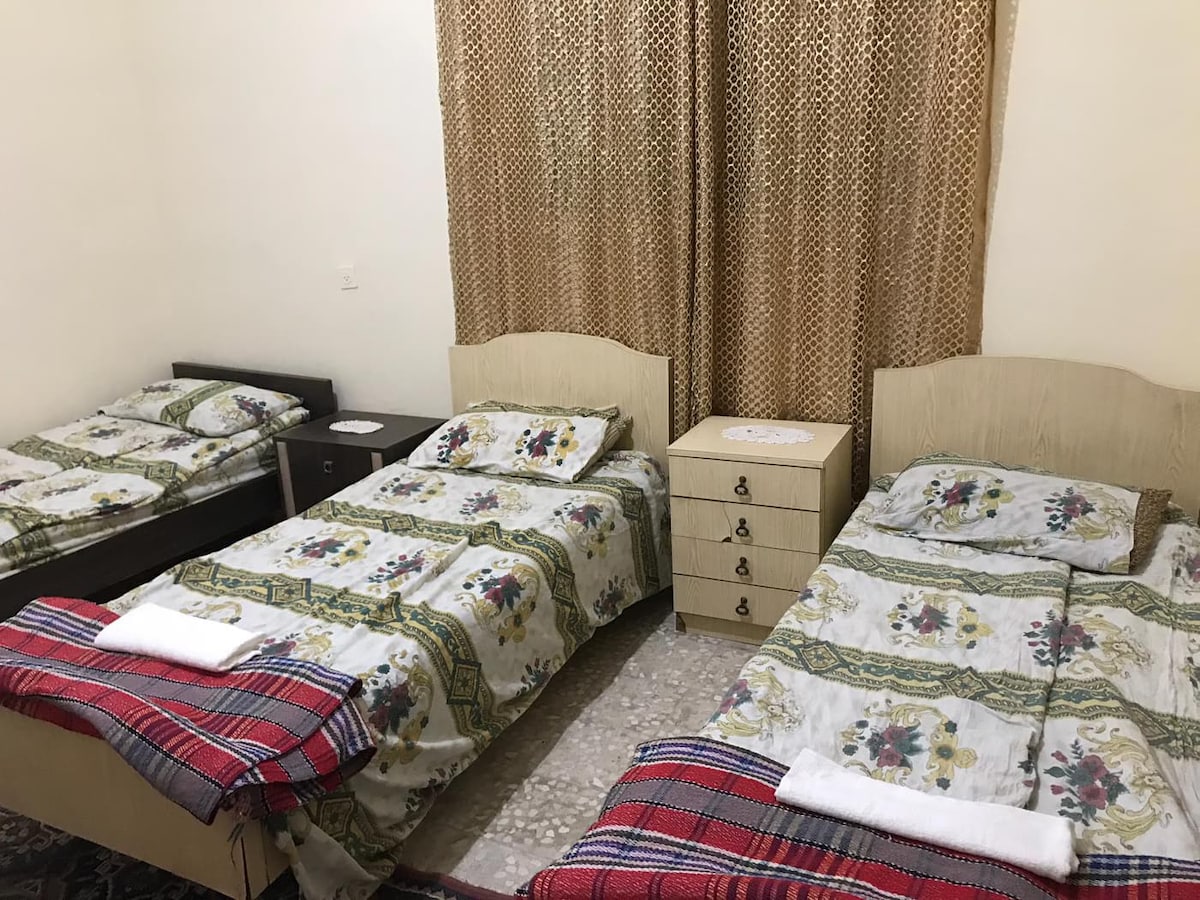 A comfortable sleeping area is furnished with three beds, each topped with patterned linens. A nightstand and a dresser are positioned nearby, and natural light filters in through a window covered with decorative curtains.