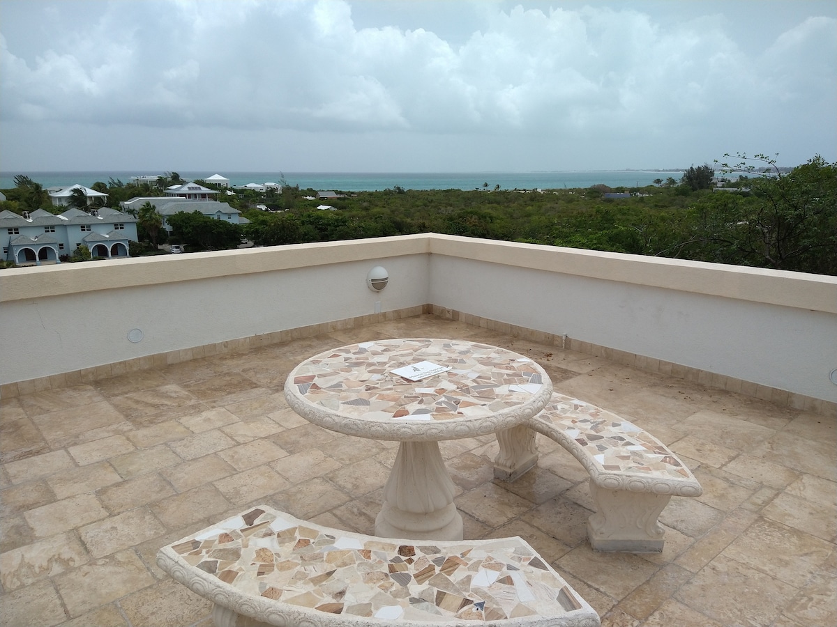 An outdoor terrace is showcased with a round stone table and two curved benches, providing ample space for seating. The area is surrounded by a tiled surface and offers a distant view of the ocean and nearby greenery under a partly cloudy sky.