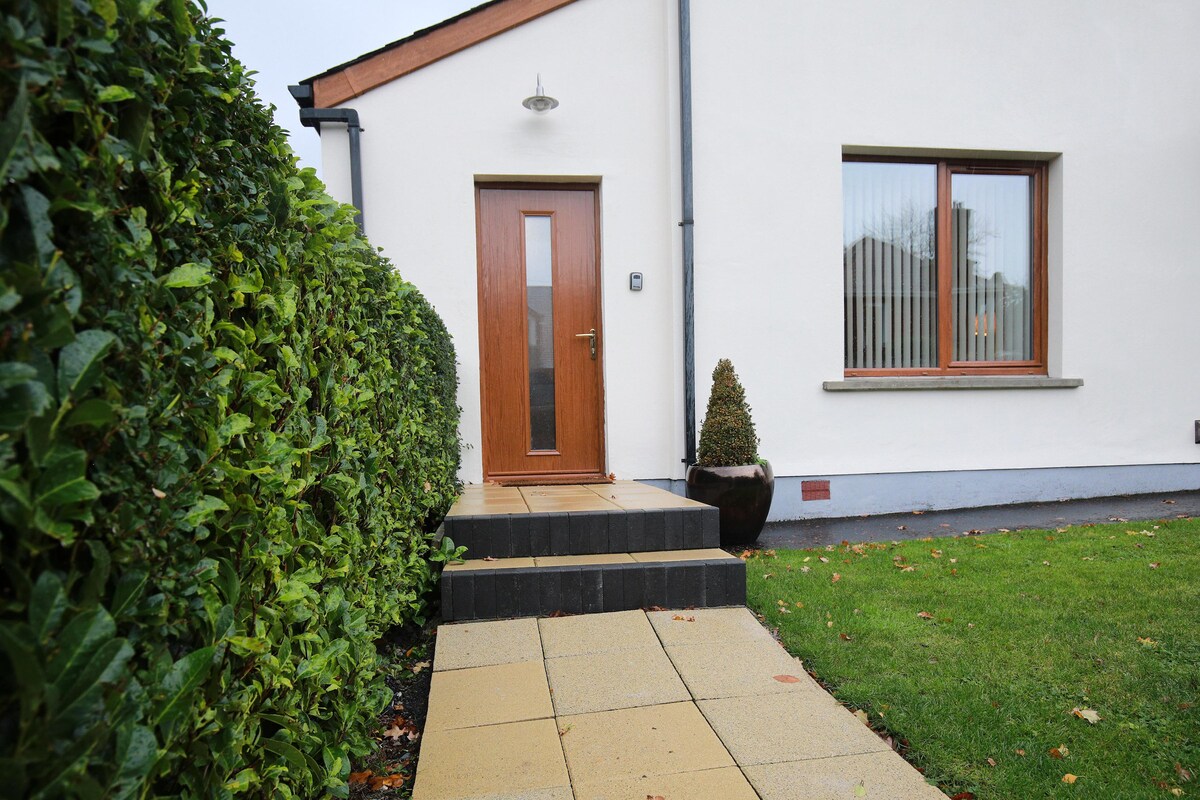 The entrance to the apartment is marked by a wooden front door with a glass panel, framed by a low hedge. A paved pathway leads up to the door, flanked by a small garden area featuring a decorative pot and well-maintained grass.