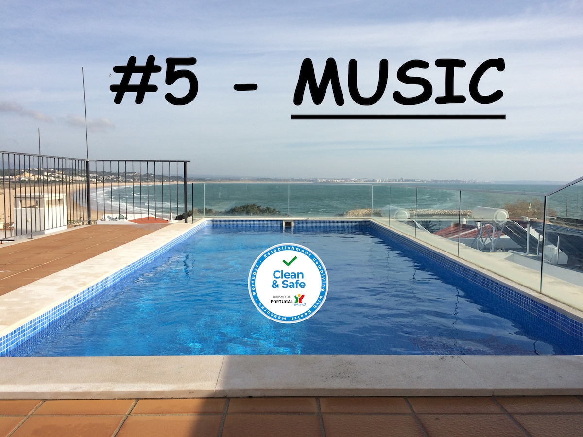 An outdoor pool is visible, showcasing clear blue water with a tiled border. The area is enclosed by glass railings, providing unobstructed views of the ocean. The sky above is bright, complementing the inviting atmosphere of relaxation.