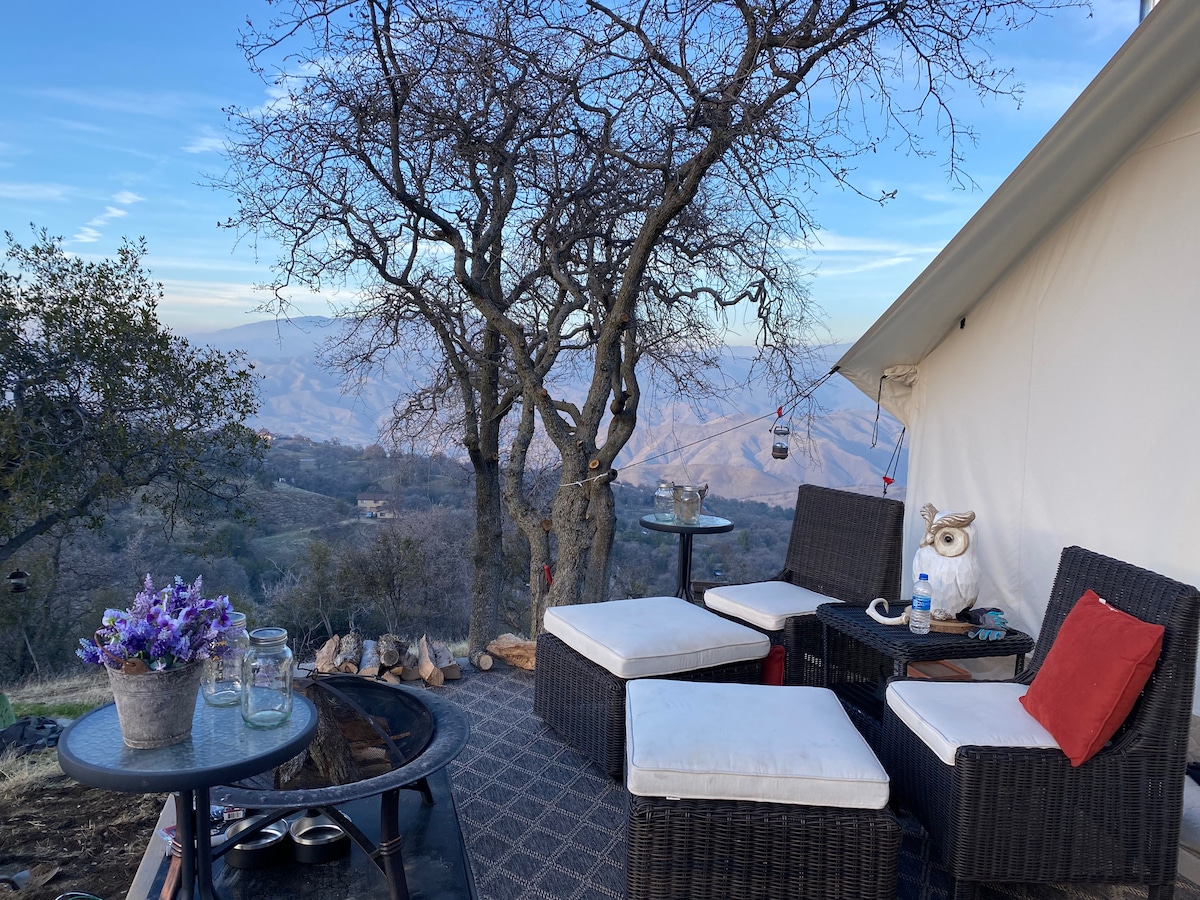 An outdoor seating area features plush white cushions on dark wicker chairs, resting beside a fire pit. Glass tables and colorful decor accompany scenic views of rolling hills and trees, while the sky transitions to twilight.