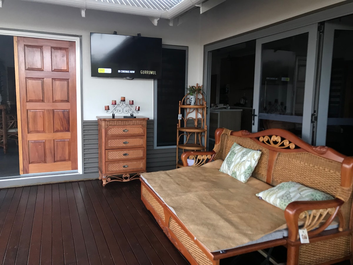 A welcoming outdoor terrace area features a comfortable wicker seating arrangement with floral cushions. A television is mounted on the wall, and a wooden cabinet displays decorative items. Glass doors lead to an interior space, enhancing the connection between indoor and outdoor living.