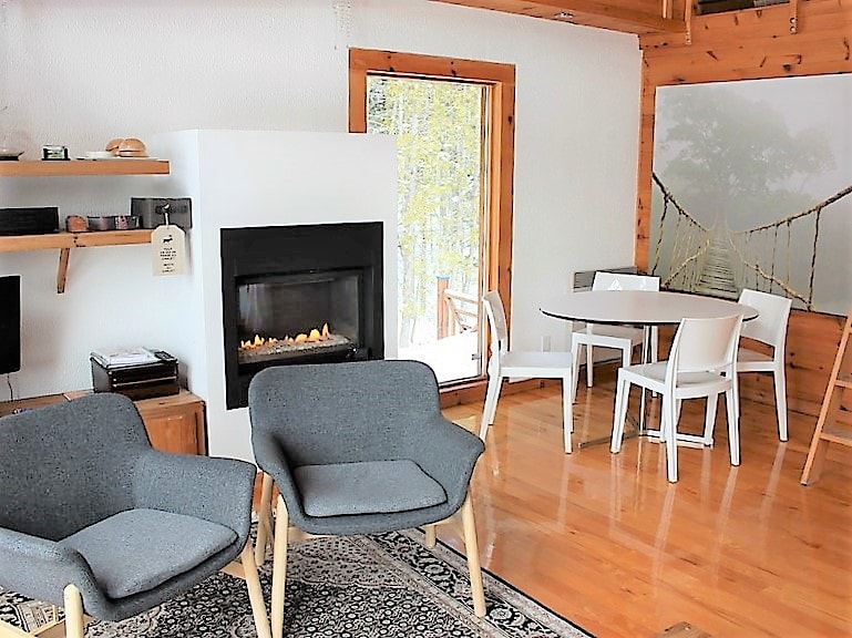 A cozy living space is highlighted by a gas fireplace, surrounded by two grey chairs and a circular dining table with white chairs. Natural light enters through a large glass door, complementing the wooden accents and polished floor.