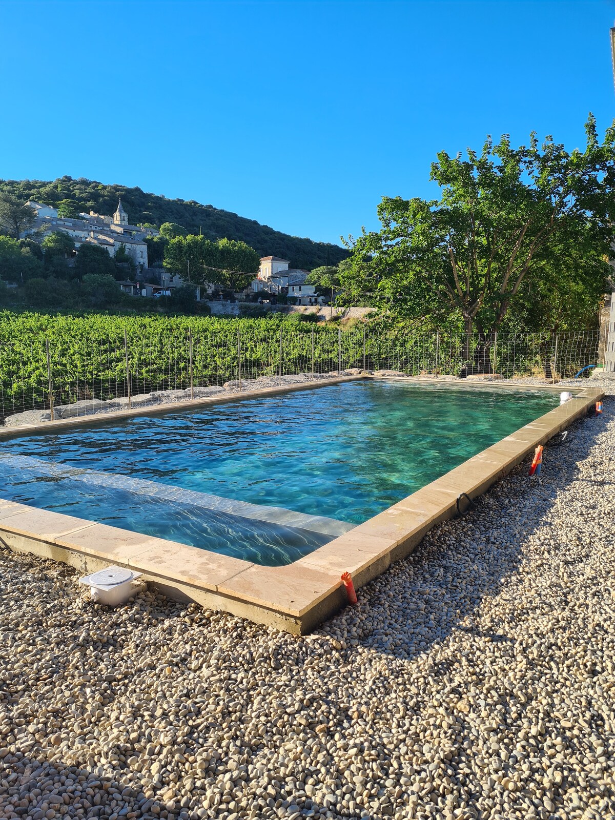 A newly constructed pool set within a gravel area, featuring clear blue water that reflects the surrounding landscape. Lush greenery and distant buildings are visible in the background, contributing to the serene outdoor environment.