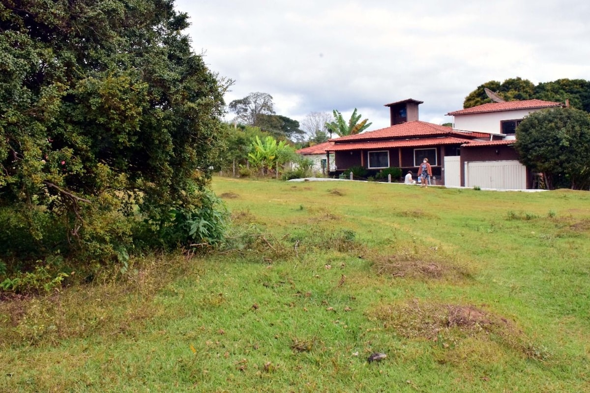 A spacious outdoor area is surrounded by greenery, featuring a well-maintained lawn. The house displays a traditional design with a red-tiled roof and large windows. A small porch adds to the inviting appeal of the property, set against a backdrop of trees and gentle hills.