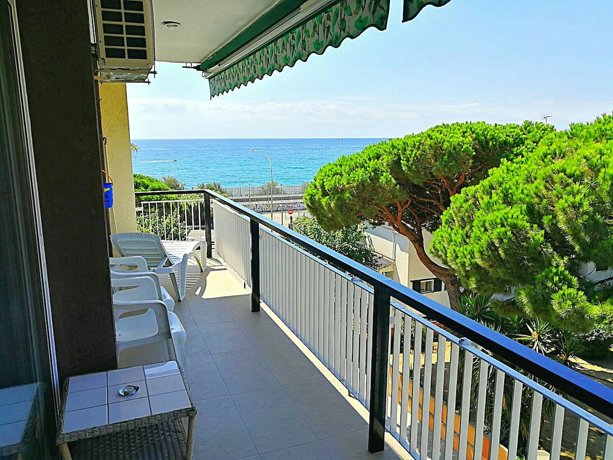 A bright balcony offers views of the sea, complemented by lush green trees. The space is furnished with several white chairs and features a shaded area, creating a relaxing environment. Sunlight streams gently across the balcony floor.