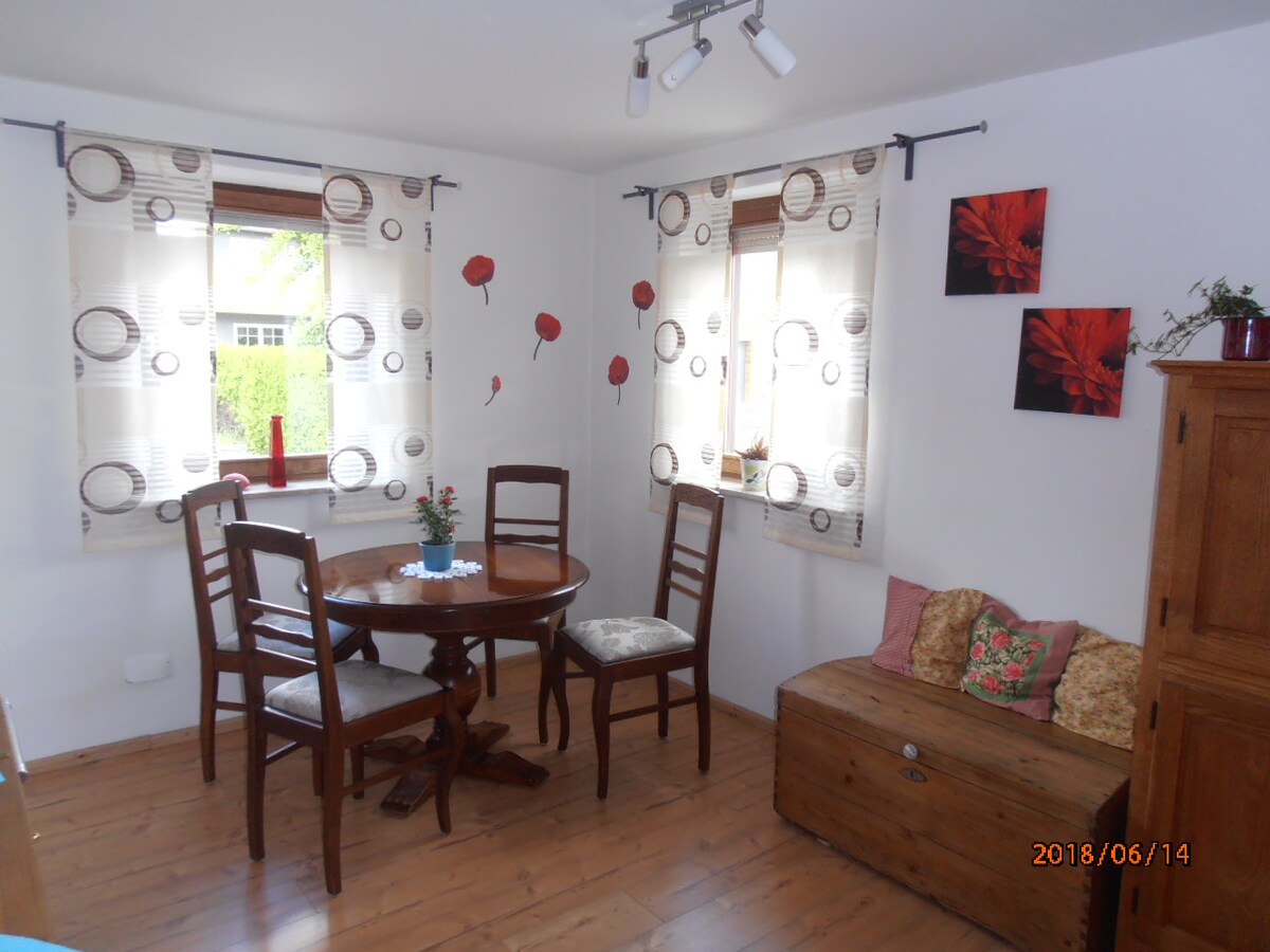 A dining area features a round wooden table surrounded by four chairs. Two windows allow natural light to fill the space, adorned with patterned curtains. A small bench with colorful cushions is positioned against one wall, complemented by decorative flower wall art.