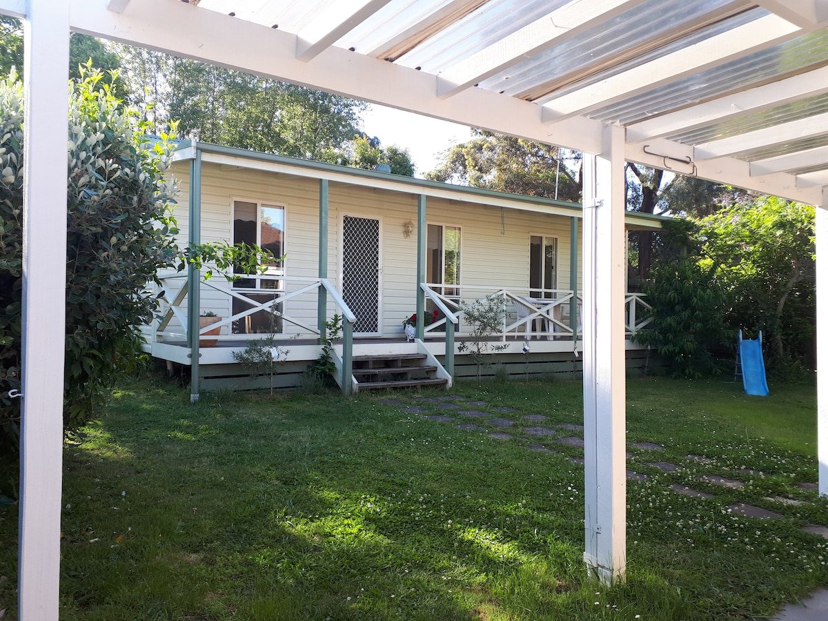 The image displays a light-colored bungalow with a covered porch, set against a backdrop of lush greenery. A gentle slope leads to the entrance, which features white railings and steps. The yard is well-kept, with soft grass and a small slide visible to the side.
