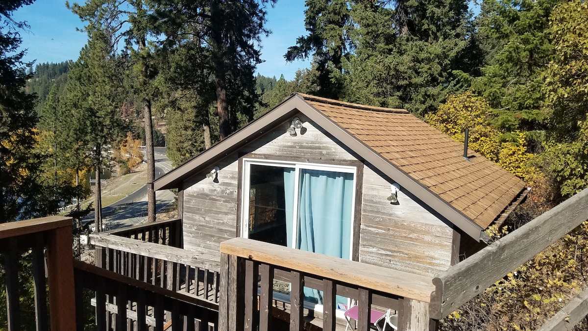 A charming cottage is nestled among trees, featuring a sloped roof and light-colored wood siding. Large windows, framed with curtains, invite natural light. A deck made of wooden planks leads up to the entrance, surrounded by vibrant foliage typical of the area.
