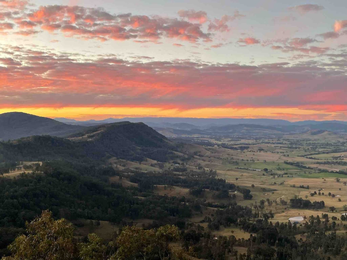 A panoramic view of rolling hills and valleys is captured at sunrise, with soft pink and orange tones illuminating the sky. Green treetops and open fields are visible below, showcasing the natural landscape of the Paterson Valley and the distant Barrington Tops.