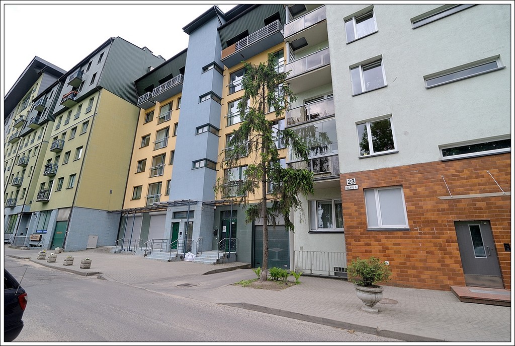 The apartment building features a modern façade with a combination of colors, including yellow and blue. Multiple stories are visible, along with several balconies. A tall tree is located in front, complementing the urban landscape, while a small planting area adds a touch of greenery along the sidewalk.
