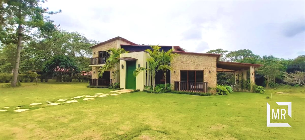 The image displays a spacious two-story cabin with stone and wood elements, set within a lush green lawn. Large windows provide natural light, while palm trees and plants frame the entrance. A covered porch with wooden accents extends the living space outdoors.