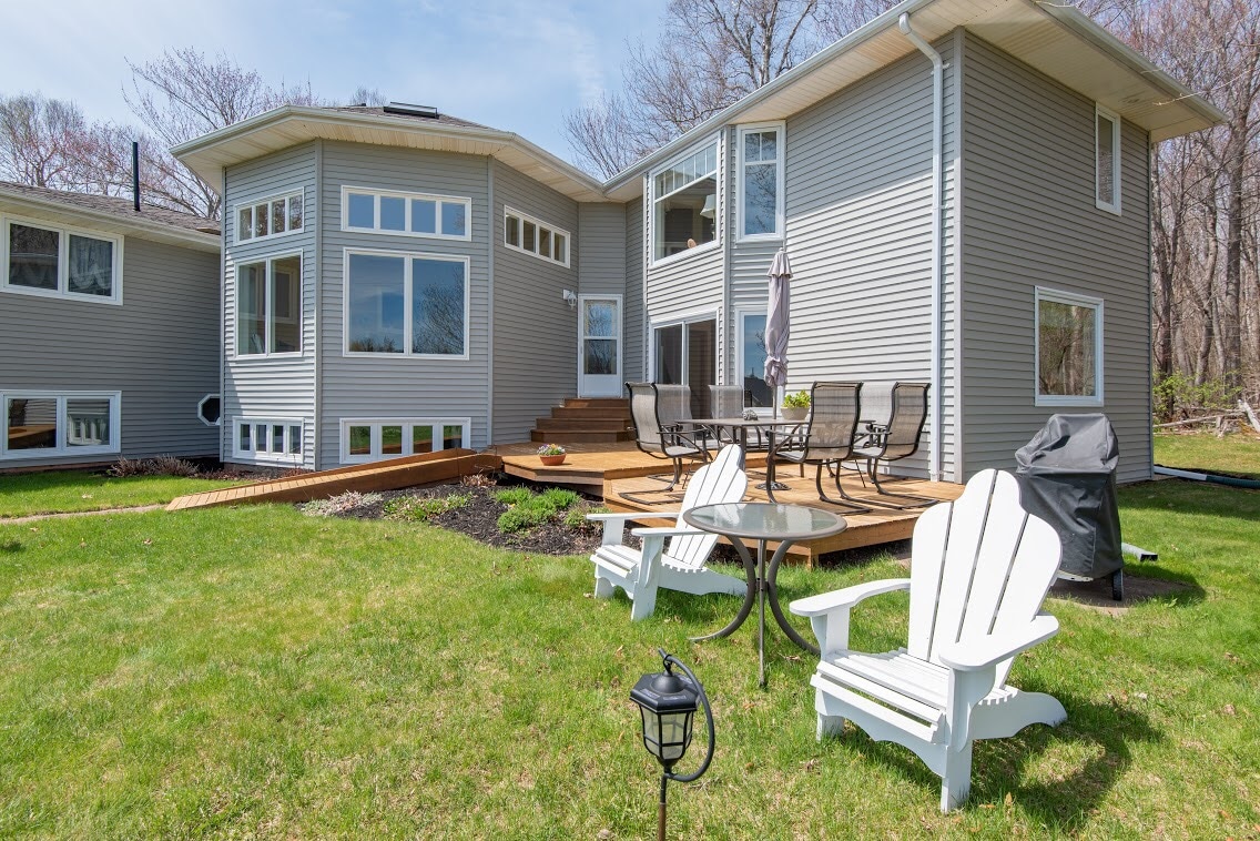The exterior of the property features a spacious deck with outdoor seating, surrounded by well-maintained grassy areas. A small table with two classic Adirondack chairs invites relaxation, while large windows reflect the natural light, enhancing the inviting atmosphere of the home.