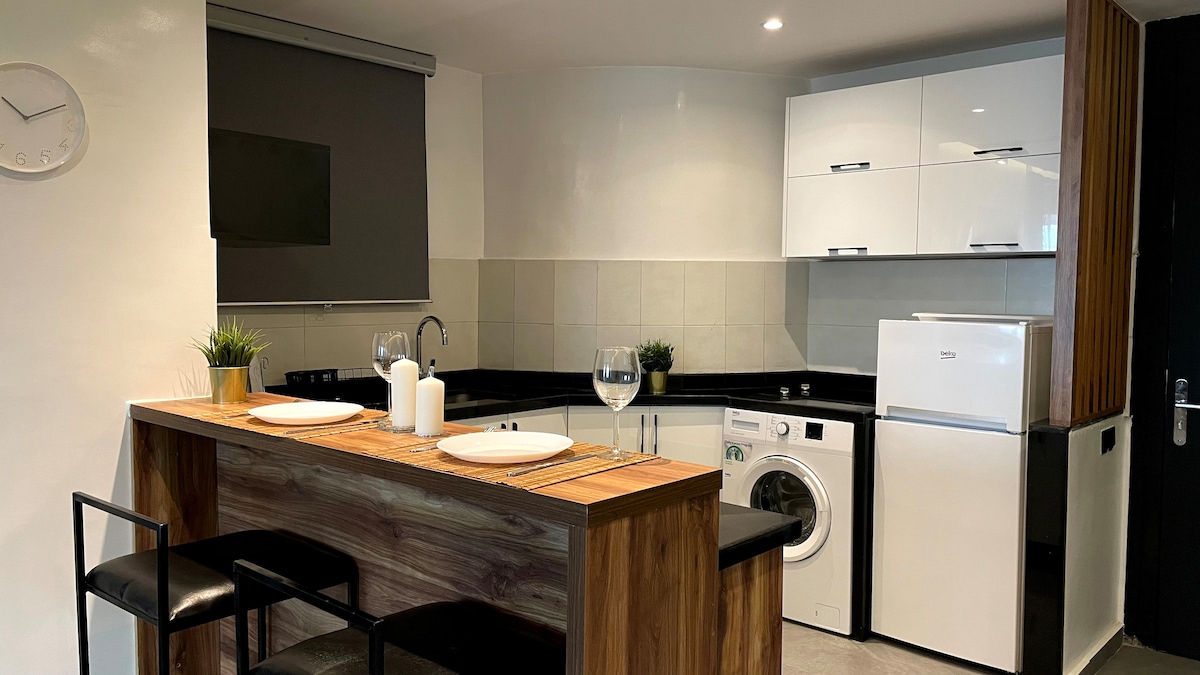 A modern kitchen space is highlighted by sleek cabinetry and a black countertop. A wooden bar with two black stools provides seating. Appliances include a washing machine, refrigerator, and sink, with decorative plants adding a touch of greenery to the ambiance.