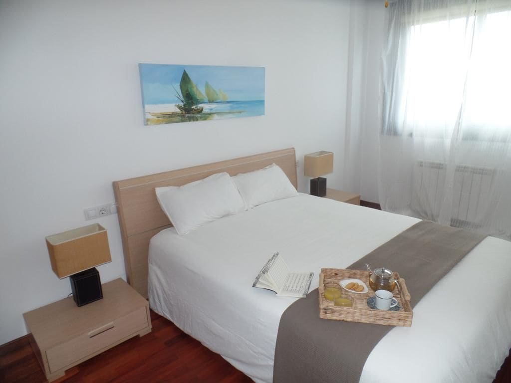 A bright bedroom features a spacious bed with white linens and a neutral-colored throw. Two bedside tables with lamps are placed on either side. A painting of sailboats is displayed above the bed, and natural light filters through sheer curtains.