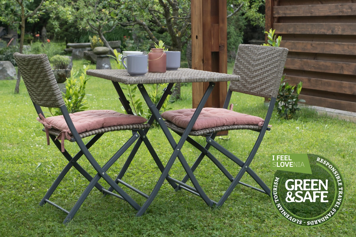 A small outdoor seating area is set in a lush green garden. A woven table is accompanied by two folding chairs, each with a cushion. Potted plants sit on the table, adding a touch of color to the inviting space surrounded by trees and stone sculptures.