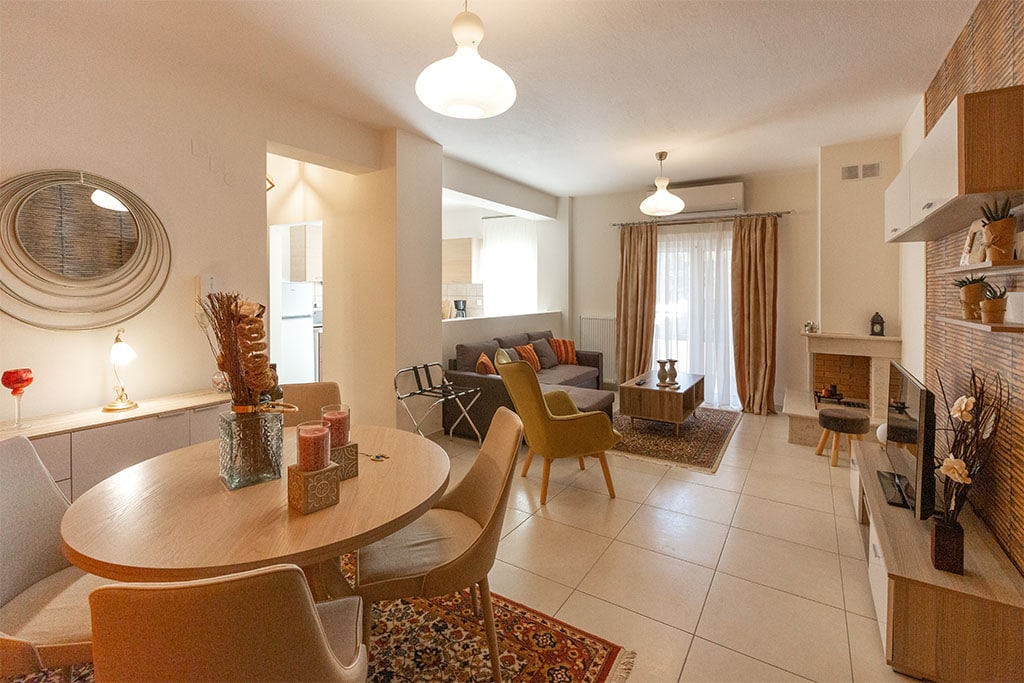 The spacious living area features a round dining table surrounded by chairs, with a cozy seating arrangement nearby. Natural light fills the room through large windows, and decorative elements, such as a wall mirror and soft lighting fixtures, complement the modern decor.