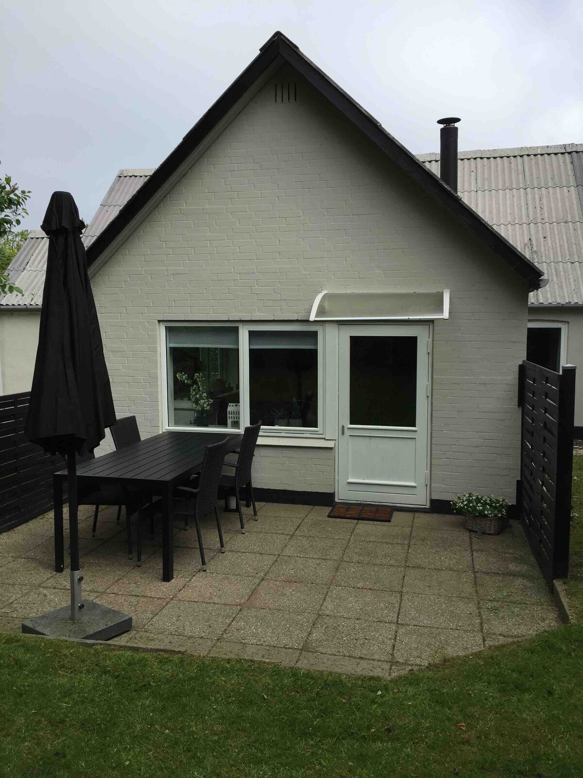 The exterior of the vacation home is presented, featuring a light grey facade with a sloped roof. A patio area with outdoor dining furniture is shown, accompanied by an umbrella for shade. A small plant is situated near the entrance, enhancing the outdoor space.