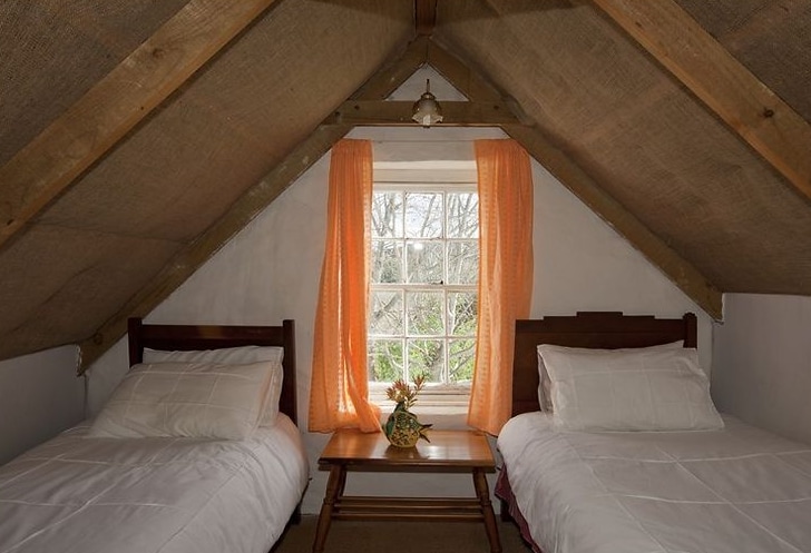 The loft sleeping area features two single beds with white linens, positioned alongside a small wooden table. A window with orange curtains allows natural light to enter, revealing greenery outside. Exposed wooden beams form the ceiling, enhancing the room's rustic charm.