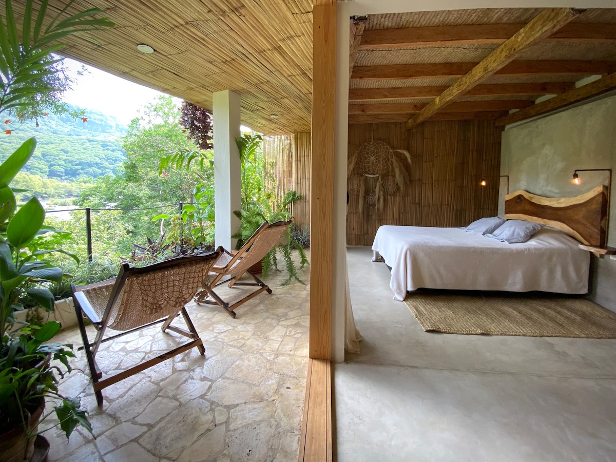A spacious living area merges with a tranquil outdoor setting, featuring a large bed and a natural stone floor. Two comfortable lounge chairs are positioned to enjoy views of the surrounding greenery. Soft lighting is provided by wall-mounted fixtures, enhancing the serene environment.