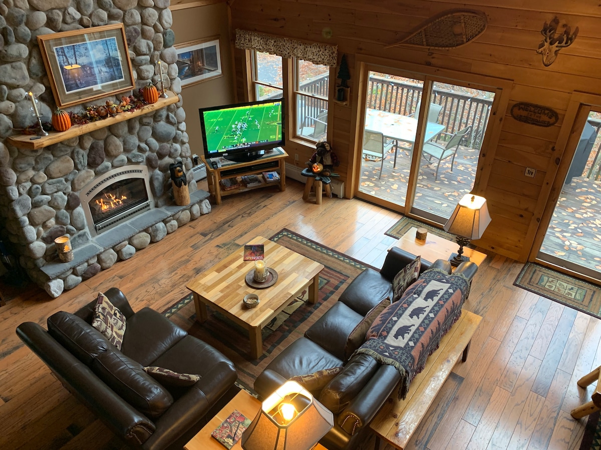 The spacious living room features a cozy seating area with dark leather sofas positioned around a wooden coffee table. A prominent stone fireplace provides warmth, while a large television faces the seating. Glass doors offer views of the outdoor deck surrounded by autumn foliage.