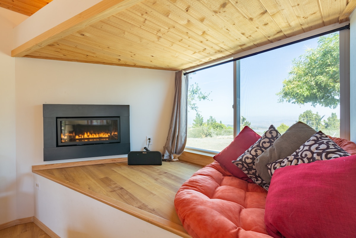 A comfortable seating area features a plush, circular couch adorned with decorative pillows. A modern fireplace is embedded in the wall below a large window that offers views of the surrounding greenery. Sunlight gently illuminates the warm wooden flooring.