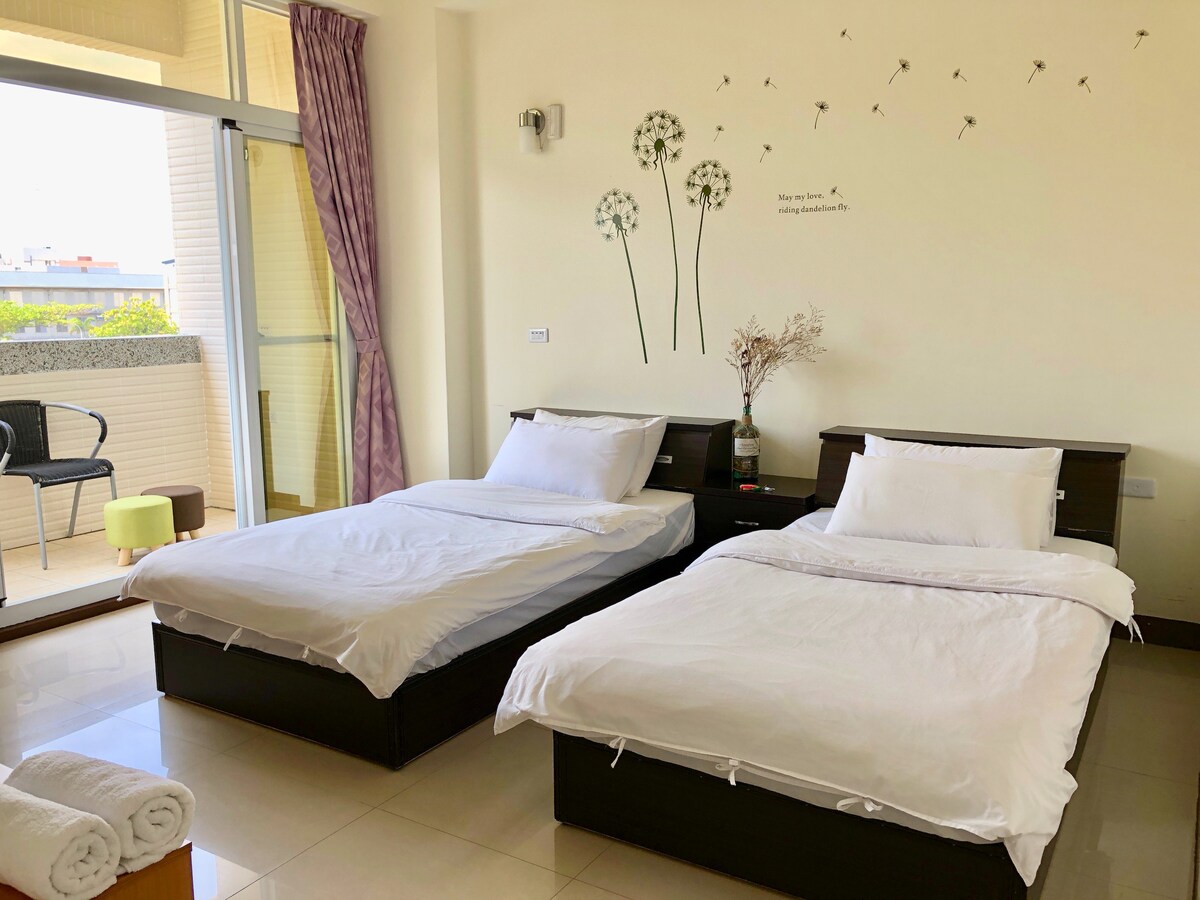 A bright room is furnished with two spacious beds positioned beside each other. A simple nightstand is located between them, topped with a decorative vase. Natural light floods the space through large glass doors, which open to a small balcony area with chairs.