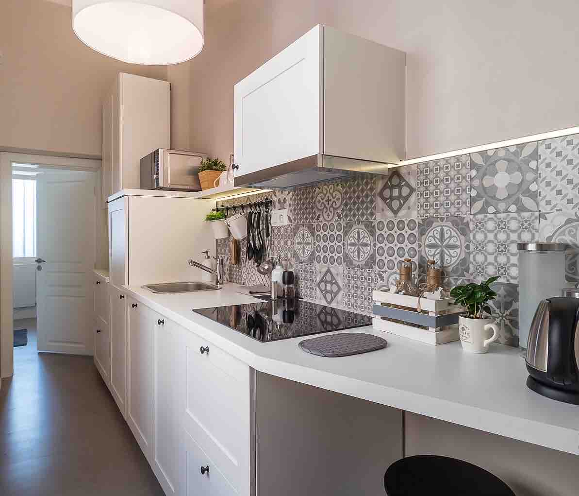 A modern kitchen features sleek white cabinetry with geometric patterned tiles as a backsplash. Cooking appliances and utensils are organized on the countertop, along with a small potted plant. A door leads to another room, enhancing the kitchen's functional layout.