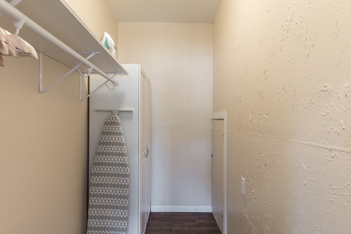 A spacious laundry area is equipped with a washer and dryer, accompanied by a full-length ironing board. Light-colored walls enhance the room's openness, while a small door is visible at the far end, potentially leading to additional storage or areas.