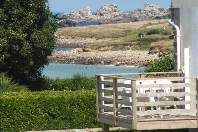 A view from a wooden terrace, showing a sandy beach and rocky coastline in the distance. Lush greenery frames the scene, with individuals enjoying the water. The clear sky enhances the bright and inviting atmosphere of this coastal location.
