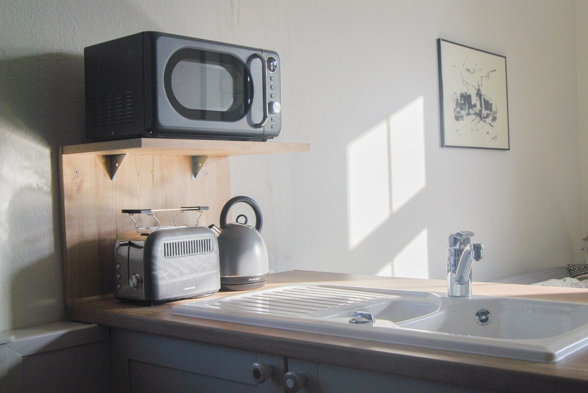 A functional kitchen area features a wooden countertop with a modern sink, complemented by a microwave, toaster, and kettle. Sunlight filters through a nearby window, illuminating the space, while a tasteful black-and-white artwork adorns the wall.