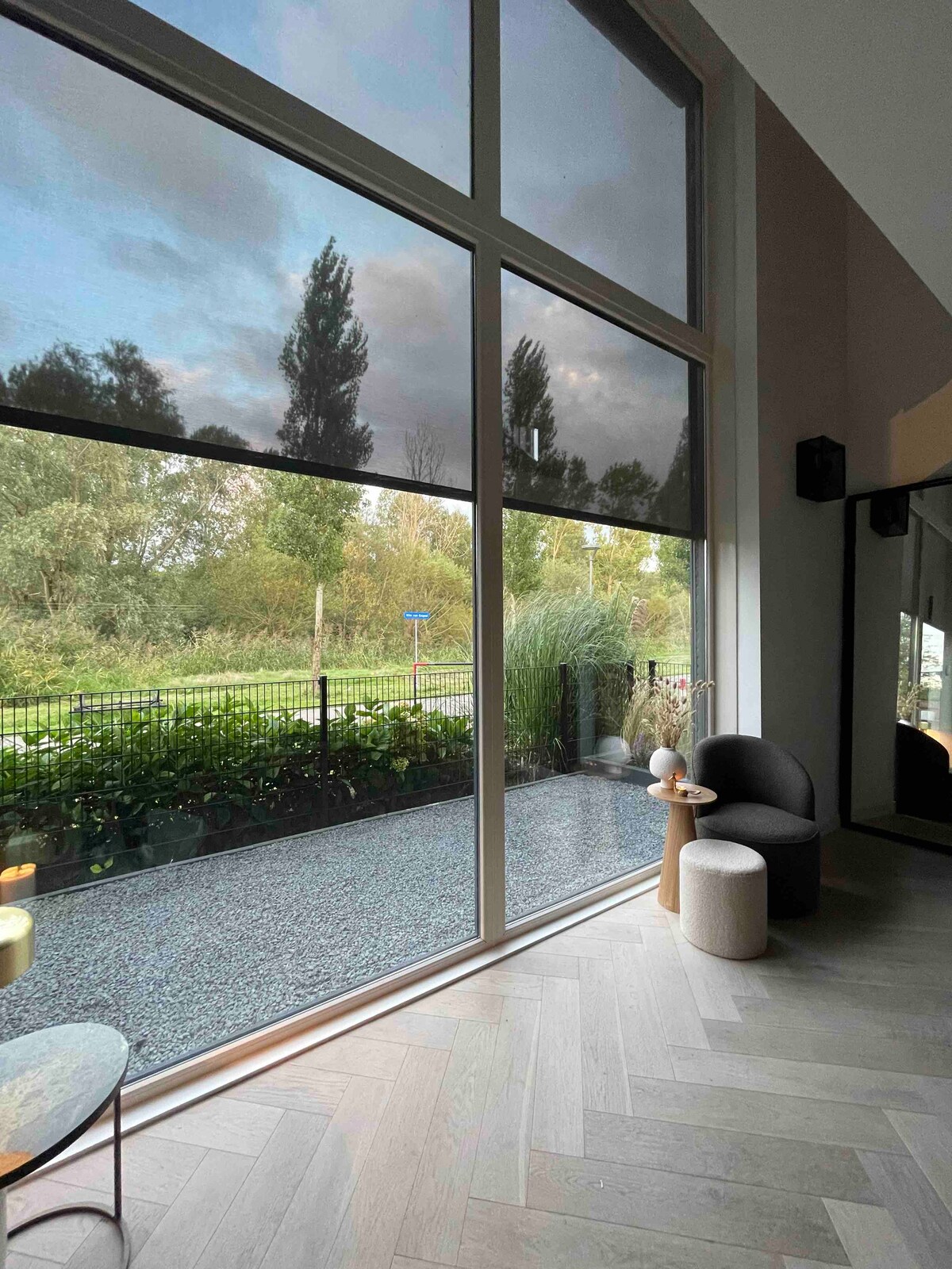 Large windows frame a view of the outdoor landscape, where green foliage and trees are visible. A cozy seating area includes a dark chair and a light-colored stool. Natural light illuminates the indoor space, enhancing the serene atmosphere.