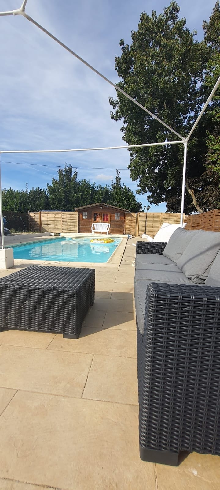 A sunlit pool area is featured, with a clear blue pool surrounded by a tiled patio. Comfortable lounge seating in dark wicker provides a casual place to relax. A wooden structure is visible in the background, with greenery enhancing the outdoor setting.