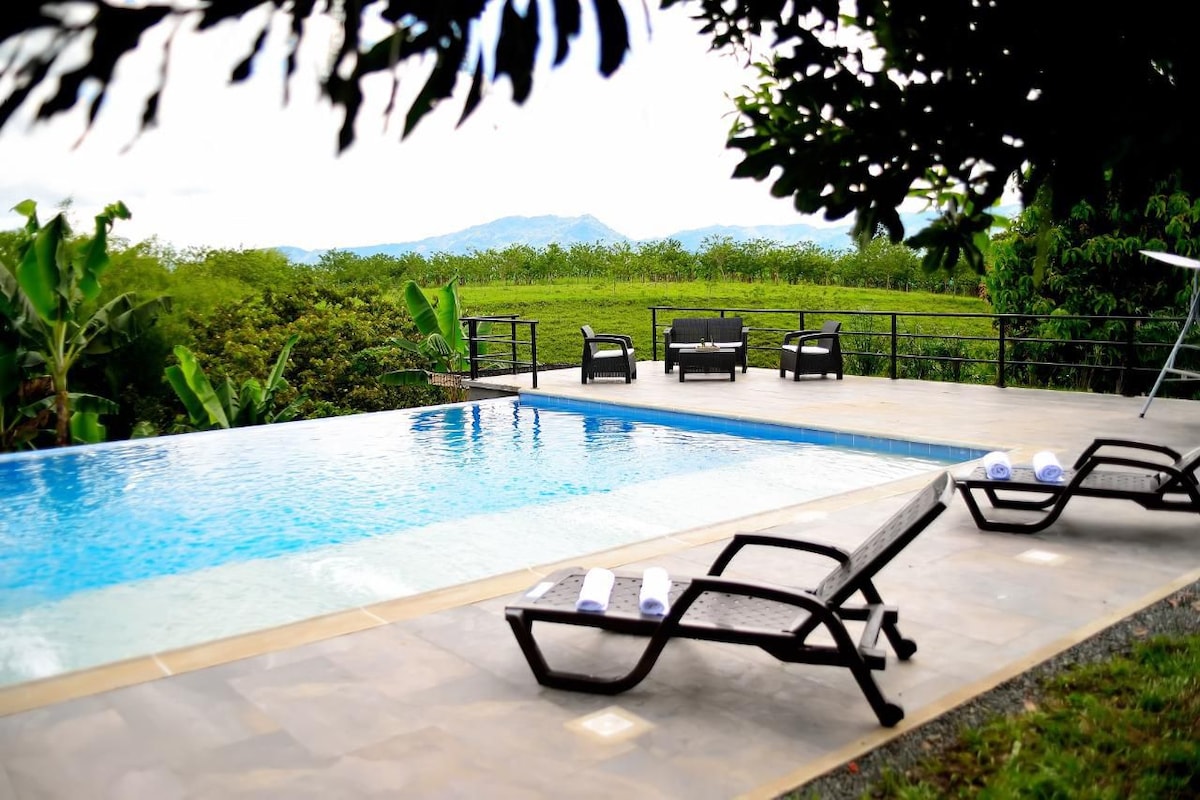 An outdoor swimming pool is situated beside a spacious deck with several lounge chairs, inviting relaxation. Lush green fields stretch into the distance, with majestic mountains visible under a cloudy sky. The area is surrounded by trees, enhancing the natural ambiance.