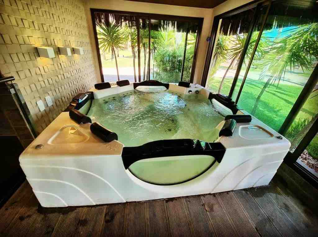 A spacious hot tub is surrounded by large glass windows, offering a view of lush greenery outside. Comfortable seats line the perimeter, with easy access for relaxation. Natural light fills the area, enhancing the calming environment of the space.